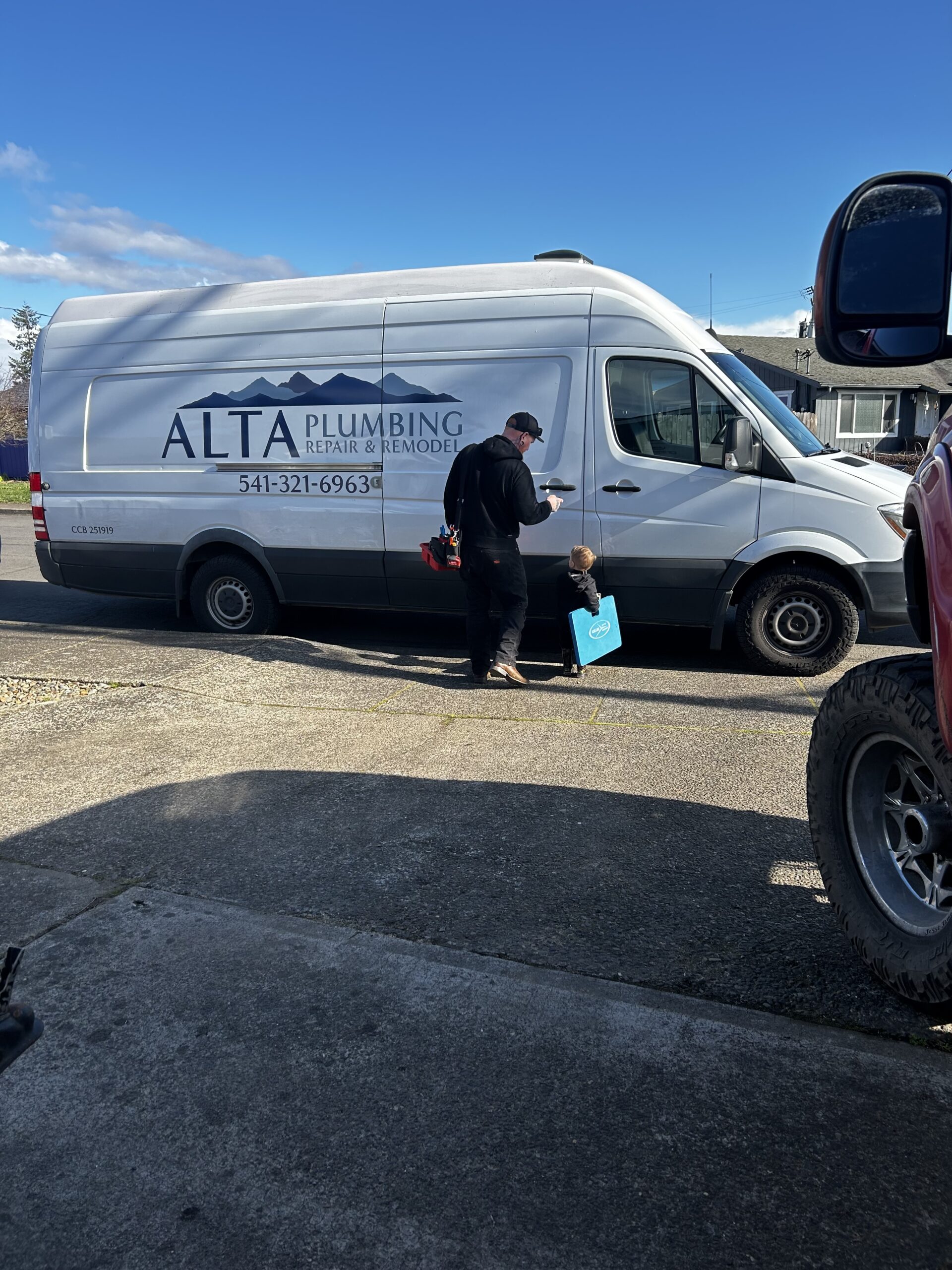 A plumber stands next to a white Alta Plumbing van on a driveway, holding tools while a young child with a blue bucket looks on—showcasing the family-friendly service highlighted in our About Us.