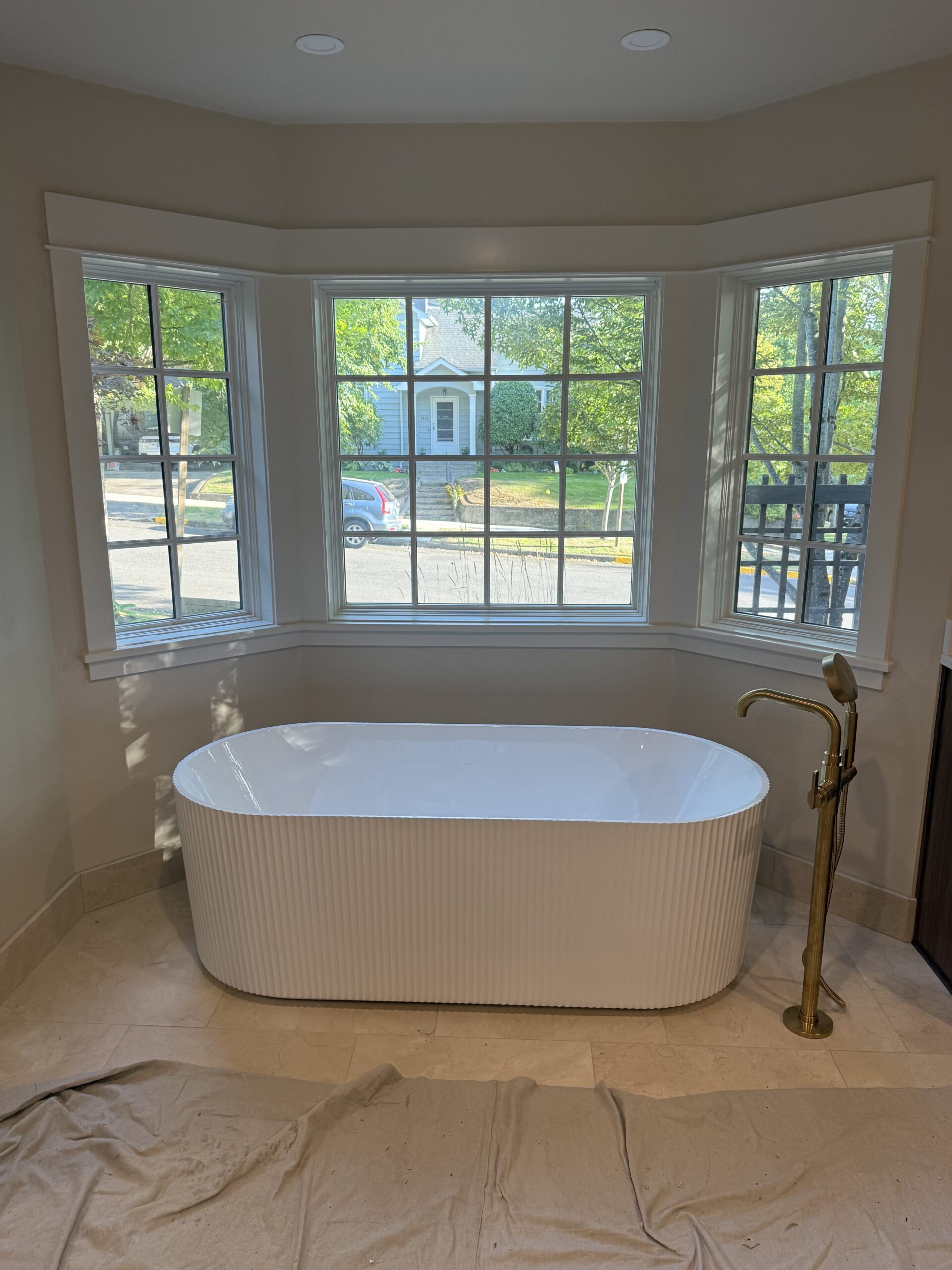 A white freestanding bathtub with ribbed sides is placed in front of three large windows in a bathroom with beige walls and flooring. A gold floor-mounted faucet stands on the right, creating a serene space that reflects our About Us design philosophy.