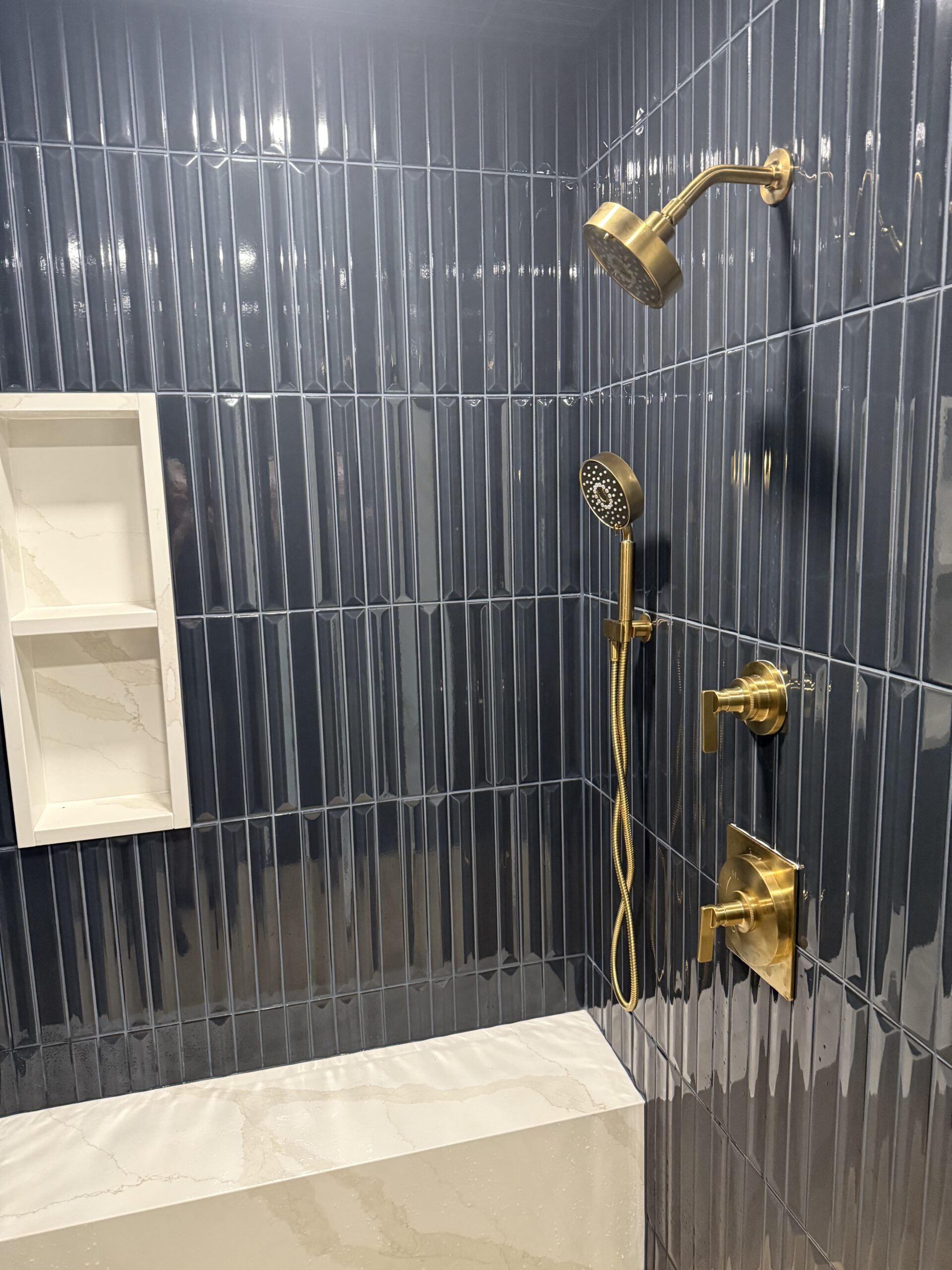 Modern shower with dark blue vertical tiles, gold shower fixtures, a handheld showerhead, and a white recessed shelf above a marble-patterned bathtub ledge—discover more design inspiration like this on our About Us page.