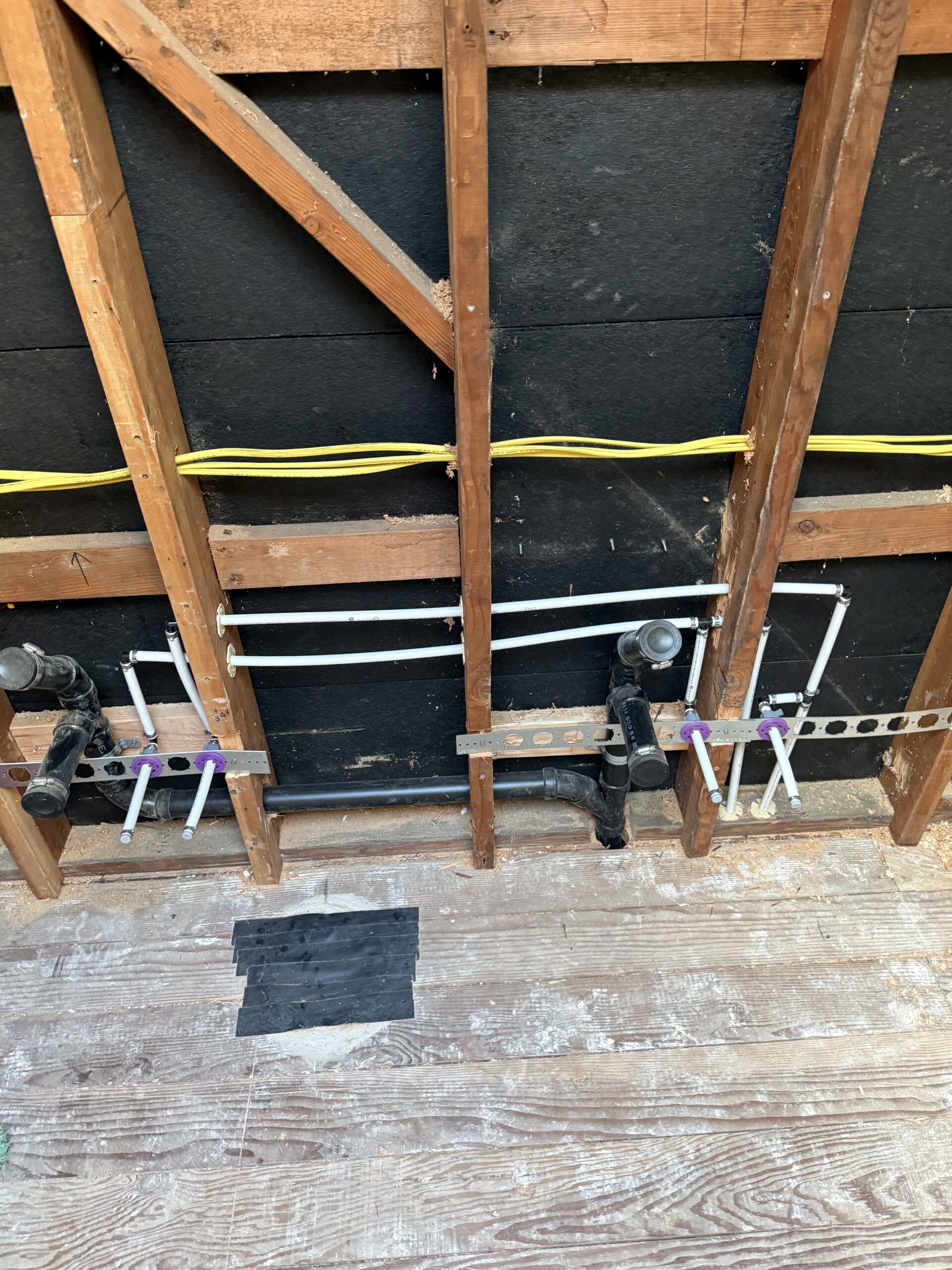 About Us: Exposed wall framing with visible plumbing pipes and electrical wiring, showing rough-in for plumbing and electrical work on an unfinished floor.