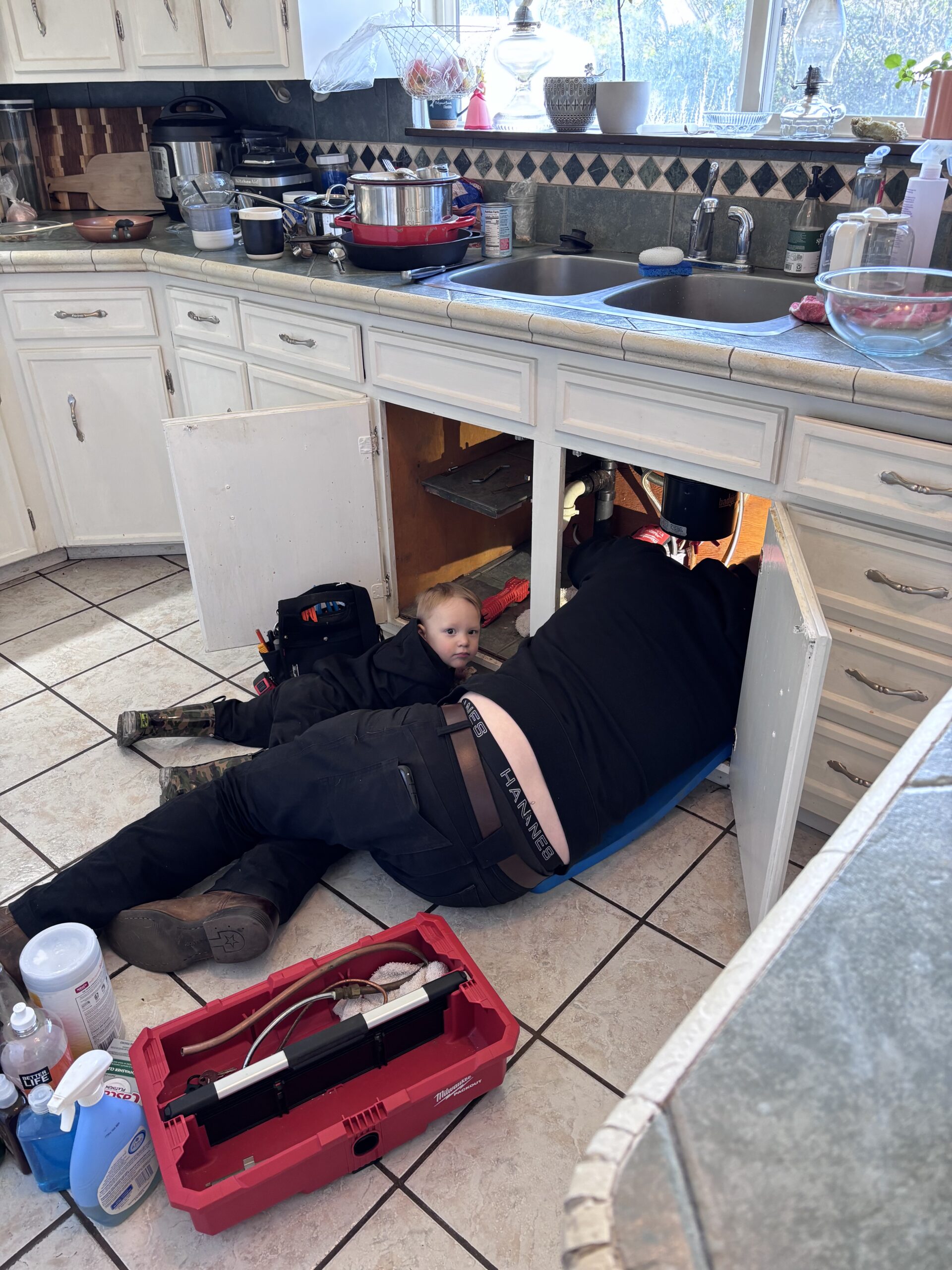 About Us: An adult and a child lie on the kitchen floor near open cabinets under the sink, surrounded by tools and cleaning supplies, as repairs are underway.