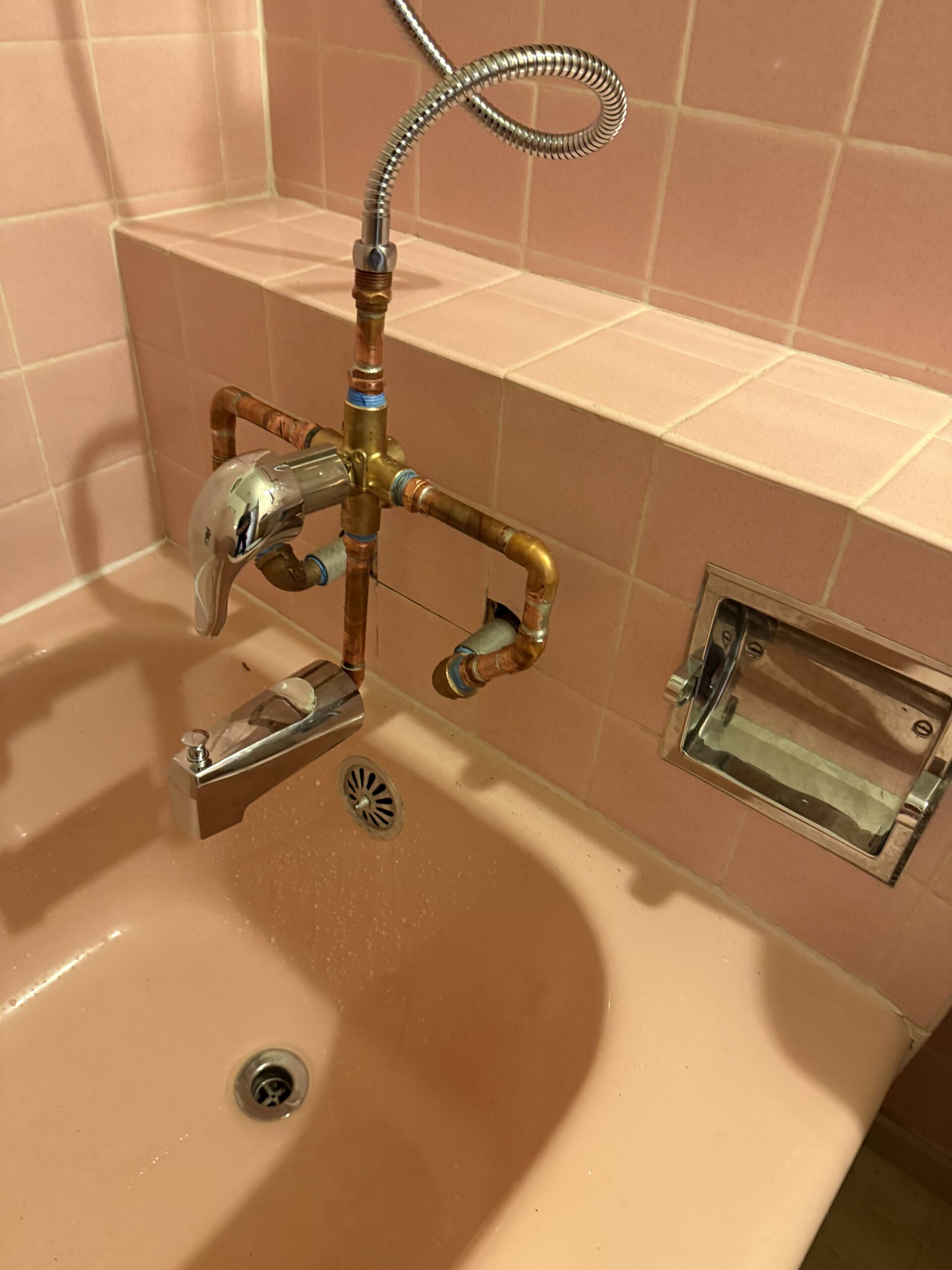 A bathtub with pink tiles, exposed copper plumbing, a modern faucet, a detachable shower hose, and a built-in soap holder offers spa-like comfort—discover more in our About Us section.