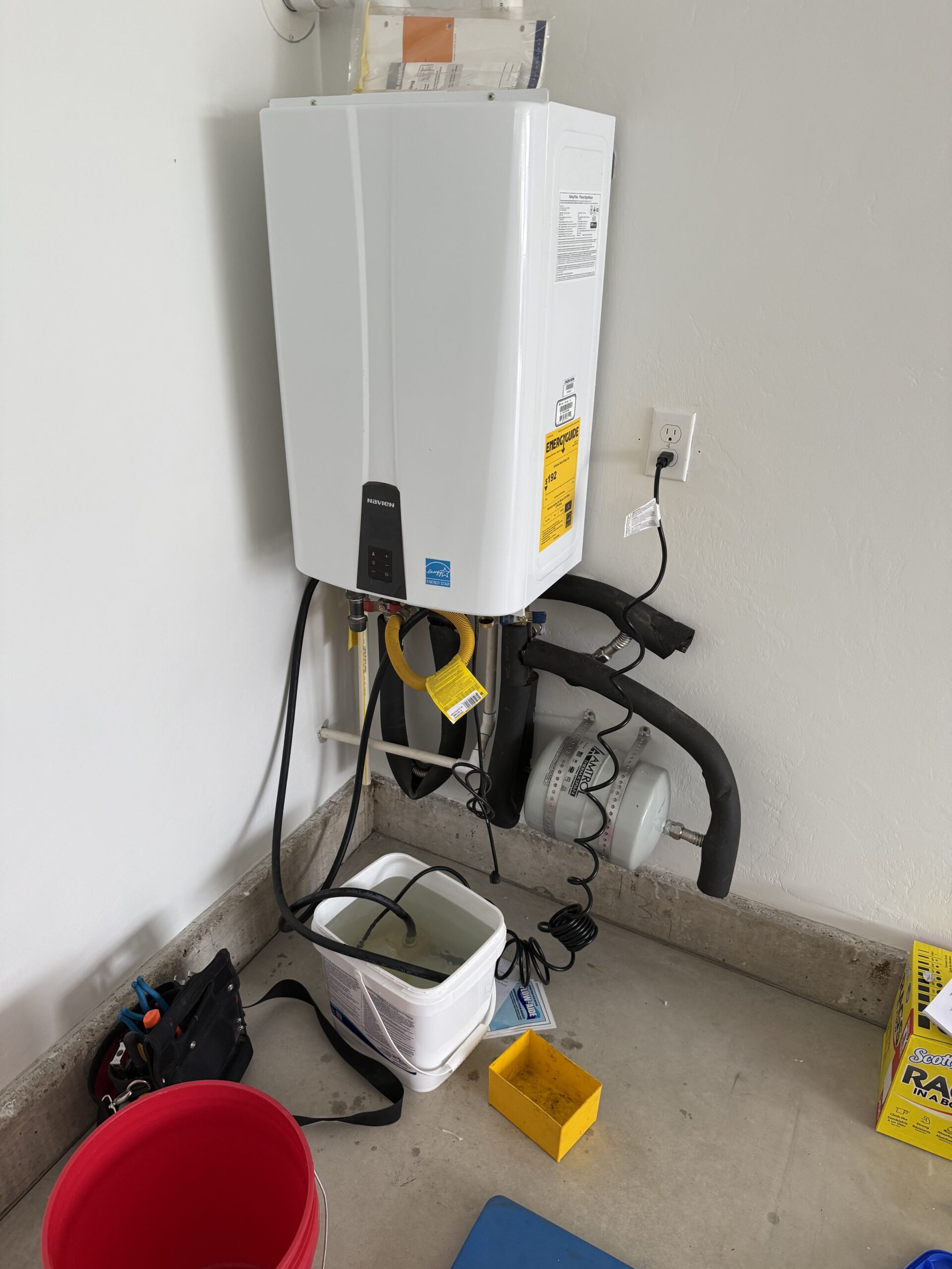A tankless water heater is mounted on a wall, with pipes, cords, a tool bag, a red bucket, and other maintenance items on the floor nearby—showcasing our expertise and commitment to quality in every About Us project.