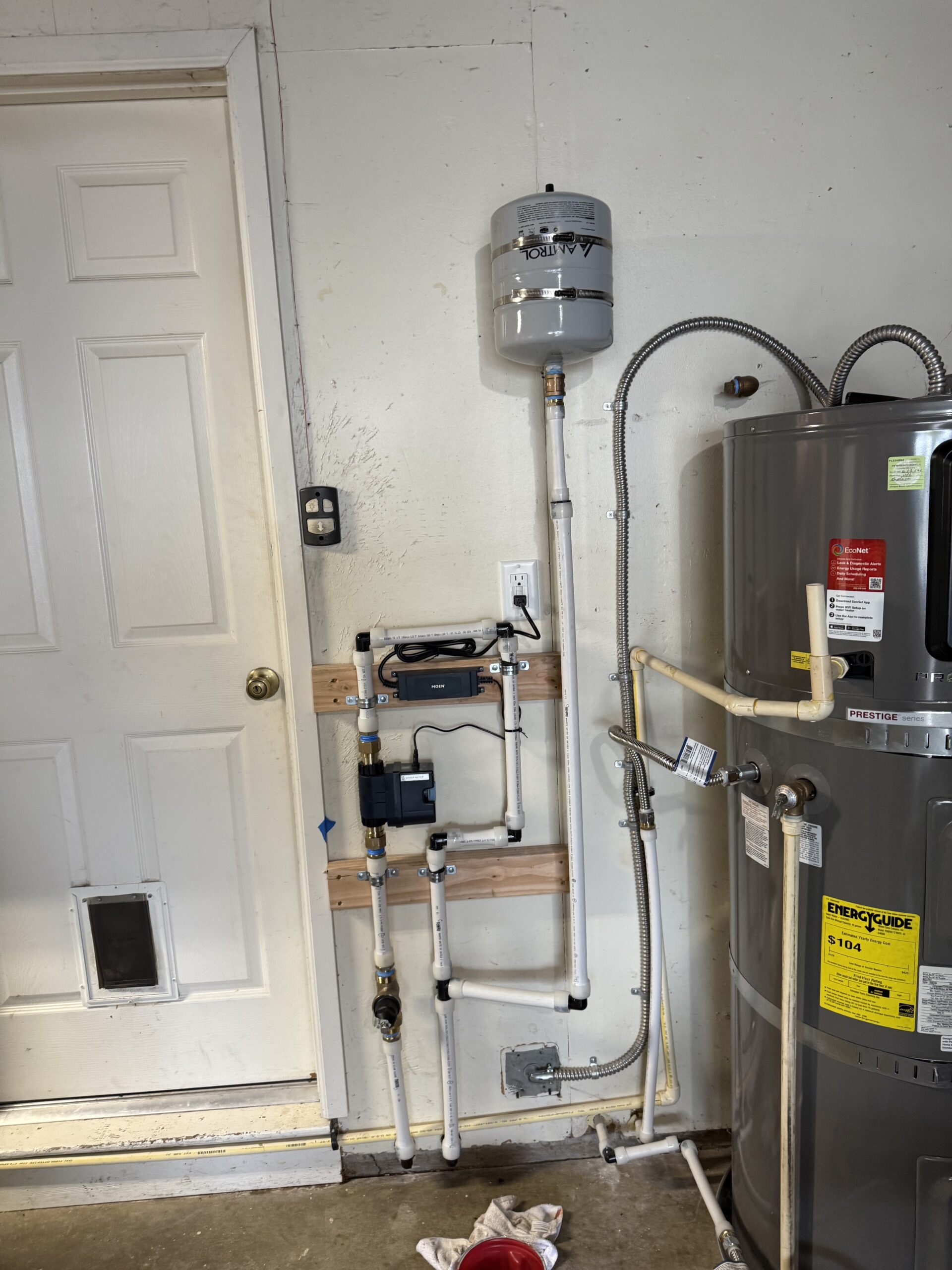 About Us: A water heater system with an expansion tank and connected plumbing is mounted on a garage wall next to a door.