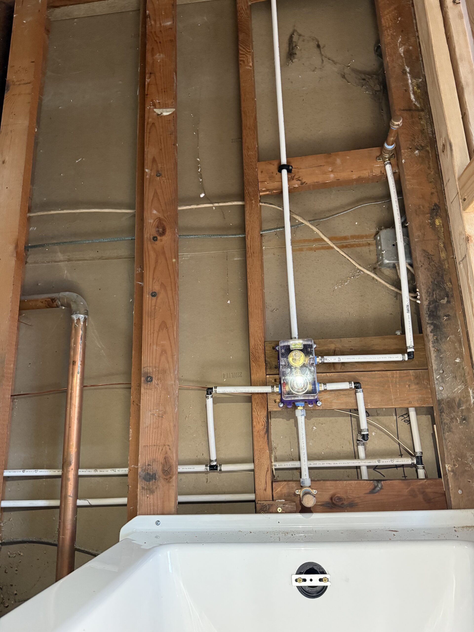 Exposed wall framing with visible copper and PVC plumbing pipes connected to a shower valve above a white bathtub—learn more about us and our craftsmanship through every detail.