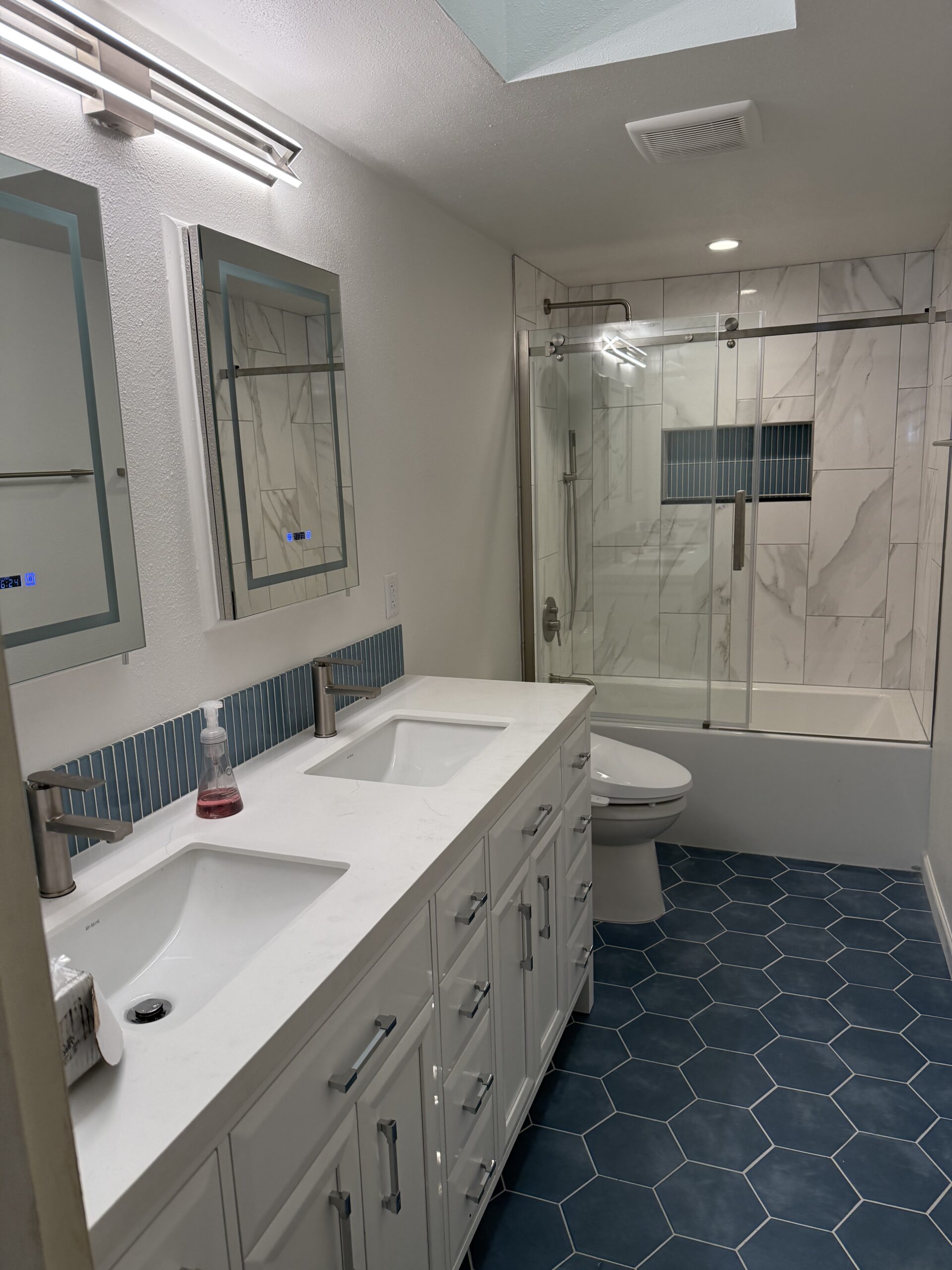Modern bathroom with double sinks, white vanity, hexagonal blue floor tiles, glass shower enclosure, and marble-tiled shower with blue accent strip—reflecting the quality and style you’ll find in every project featured in our About Us.